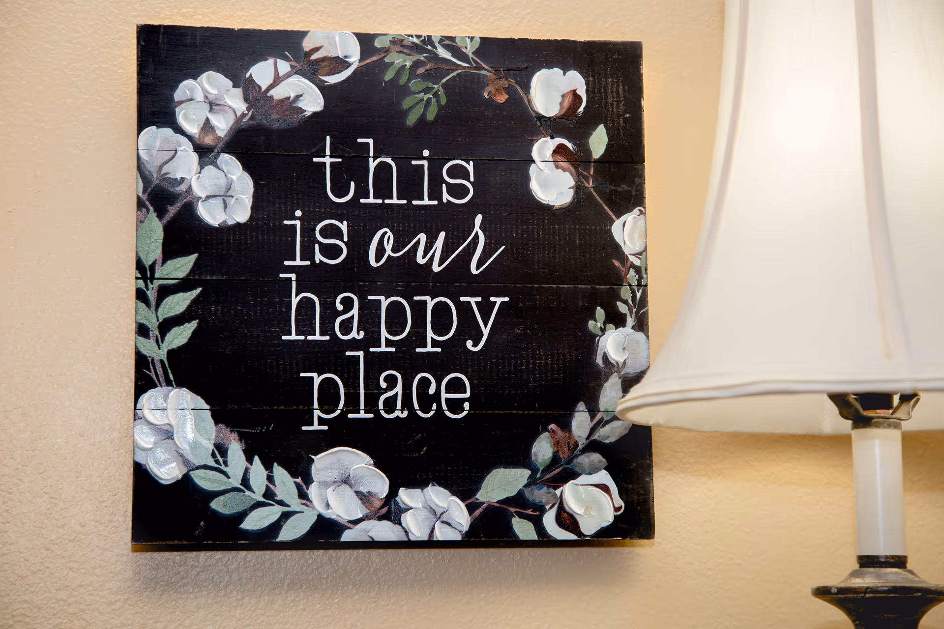 A decorative wall sign with a floral wreath design and the text 'this is our happy place' next to a white lampshade on a beige wall.