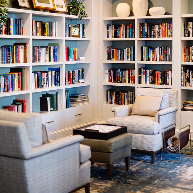 Cozy reading nook with two upholstered armchairs and a small ottoman in front of built-in white bookshelves filled with books and decorative items. A magazine rack is placed beside one of the chairs on a patterned carpeted floor.