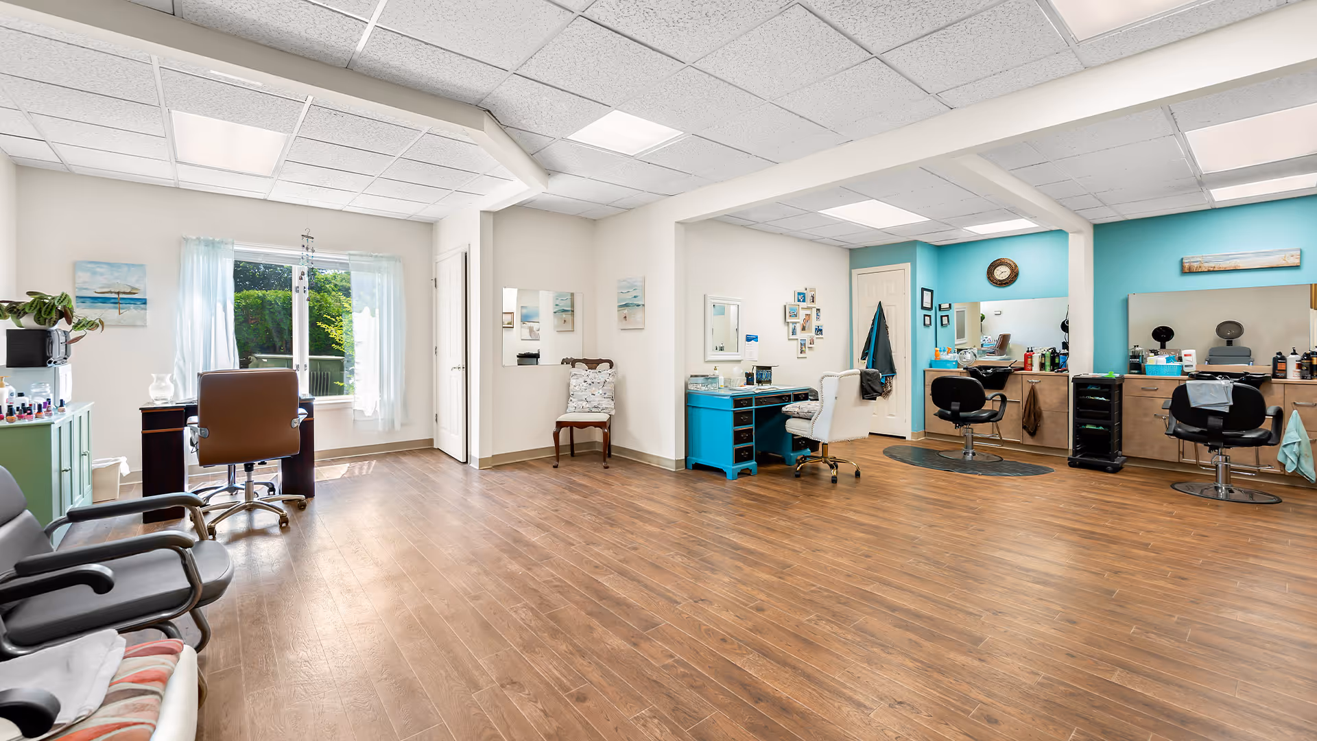 A spacious salon area in a senior living facility with wooden flooring, multiple styling chairs, mirrors, and a turquoise accent wall. There is a window with sheer curtains letting in natural light, and various salon equipment and decor are visible.