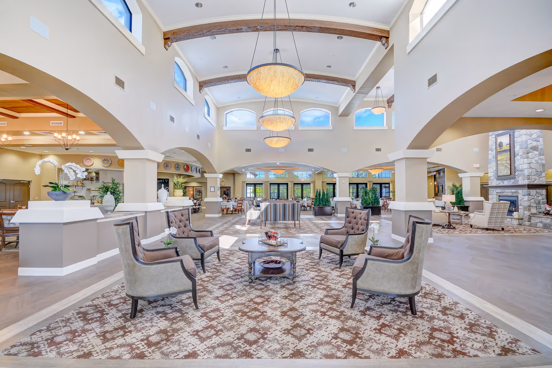 Spacious and well-lit senior living facility common area with high ceilings and large windows. The room features a seating arrangement with four cushioned armchairs around a coffee table on a patterned carpet. There are decorative plants and vases on ledges, multiple hanging light fixtures, and a stone fireplace on the right side. In the background, there are dining tables and chairs near large glass doors.