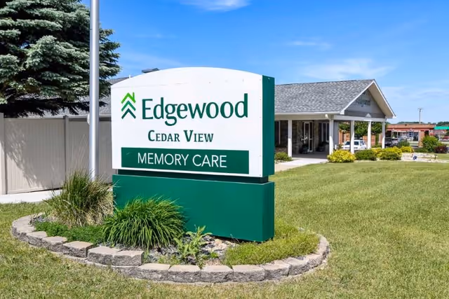 Outdoor view of the Edgewood Cedar View Memory Care facility sign surrounded by a small landscaped area with grass and plants, with the building and a covered entrance visible in the background under a clear blue sky.