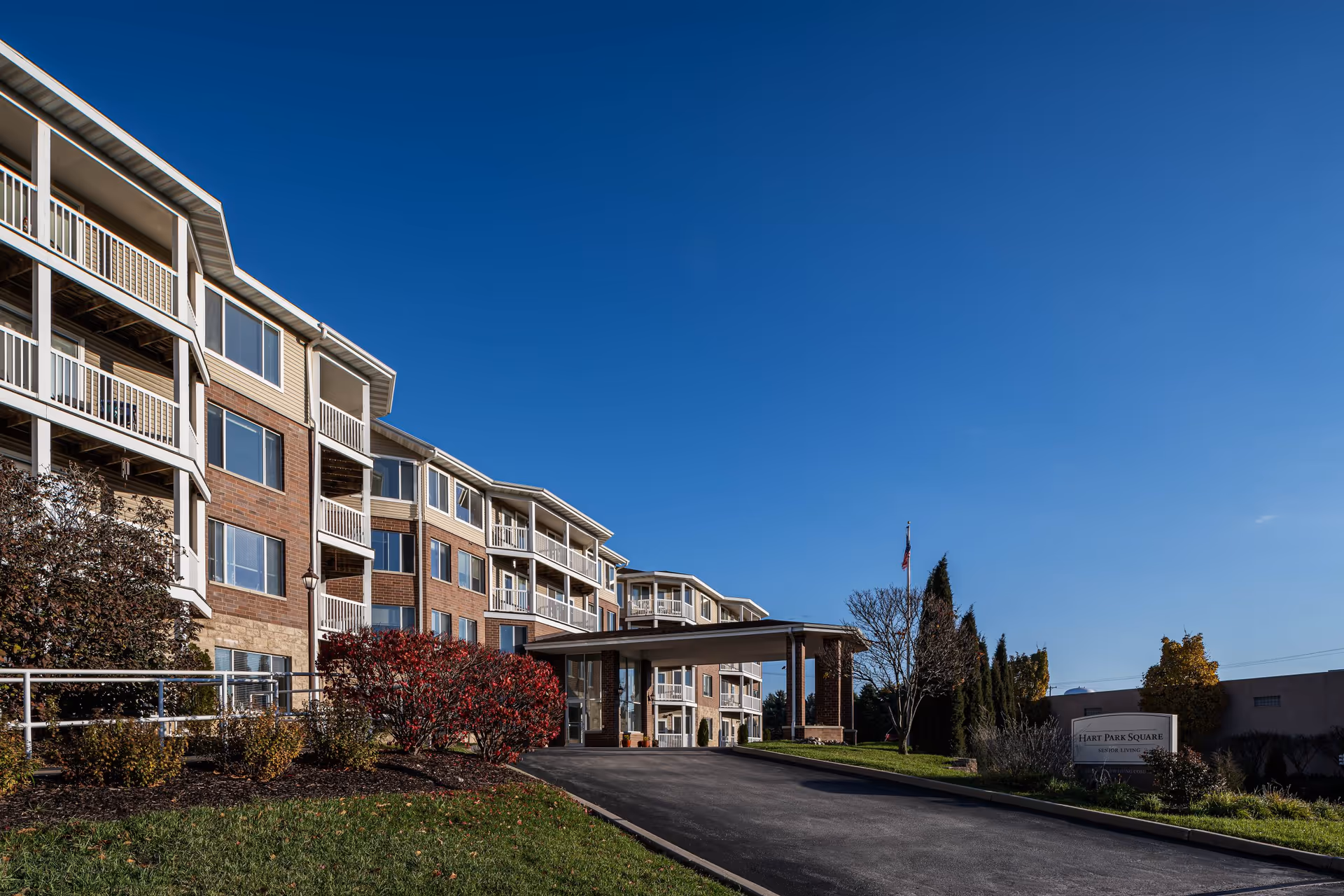 Exterior view of Hart Park Square Senior Living facility showing a multi-story building with balconies, a covered entrance, landscaped bushes, and a clear blue sky.