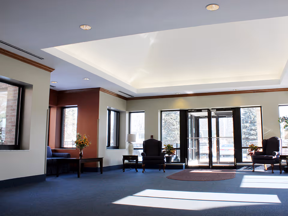 A spacious and well-lit lobby area with large windows and glass doors allowing natural light to enter. The room features blue carpet flooring, beige walls with a maroon accent wall, and a white ceiling with recessed lighting. There are several armchairs, side tables with lamps, and potted plants arranged around the space.