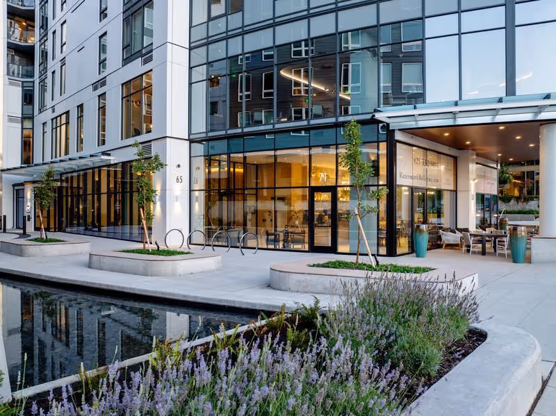 Modern multi-story building with large glass windows and an outdoor seating area. The foreground features a landscaped area with purple flowering plants and a reflecting water feature. The building has a clean, contemporary design with trees planted along the sidewalk.