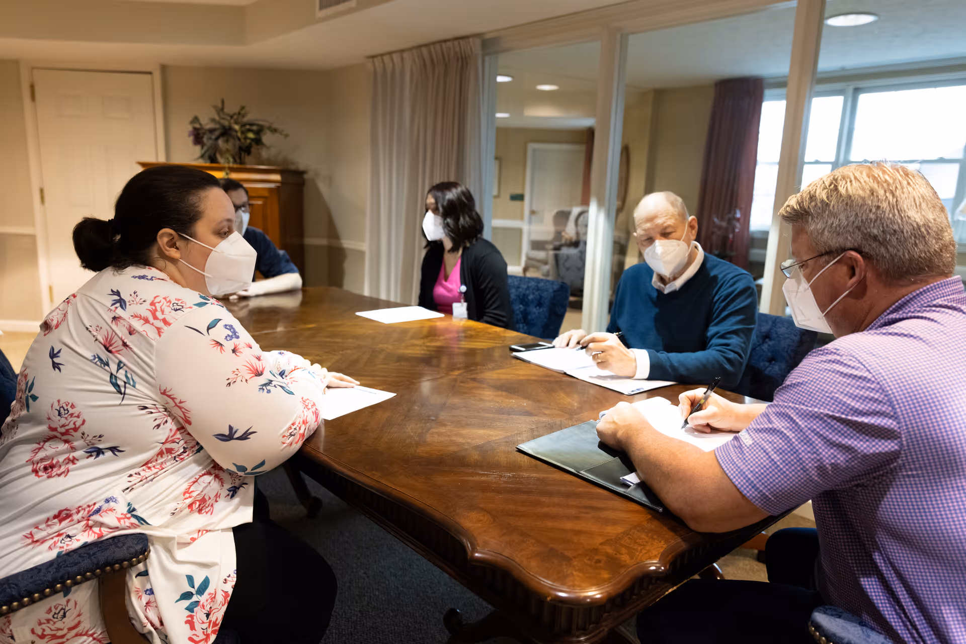 Five people wearing face masks sitting around a large wooden table in a meeting room, engaged in discussion and taking notes.