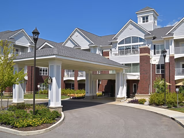 Front exterior view of Irene Woods Assisted Living facility showing a large covered entrance with white pillars, brick and white siding building, landscaped greenery, and a clear blue sky.