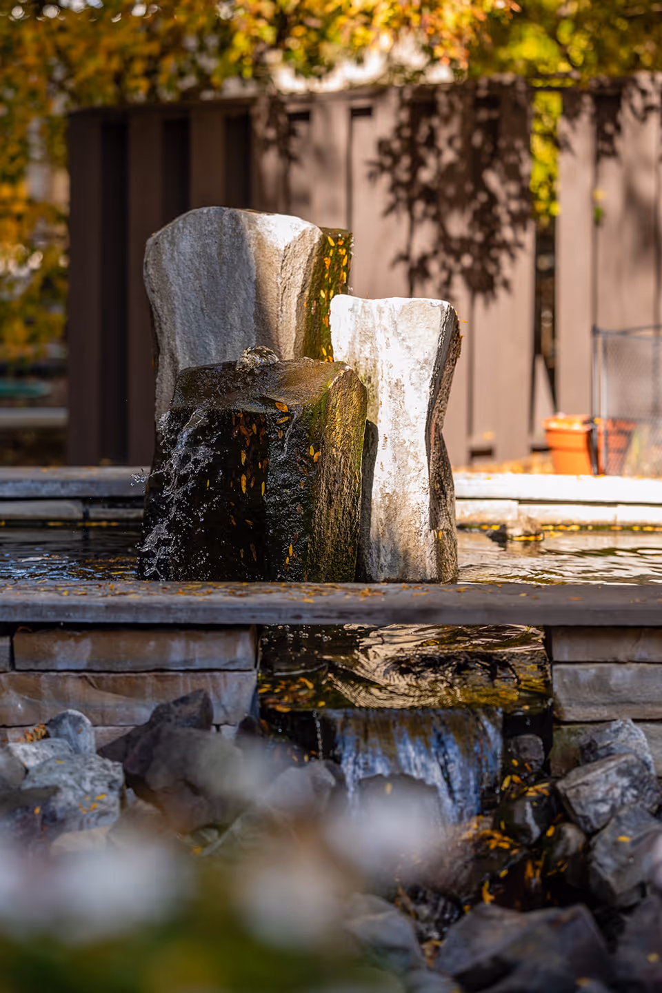A close-up view of a stone water fountain with water flowing over dark and light textured rocks, surrounded by a stone basin. In the background, there is a brown wooden fence and some autumn foliage with yellow leaves.