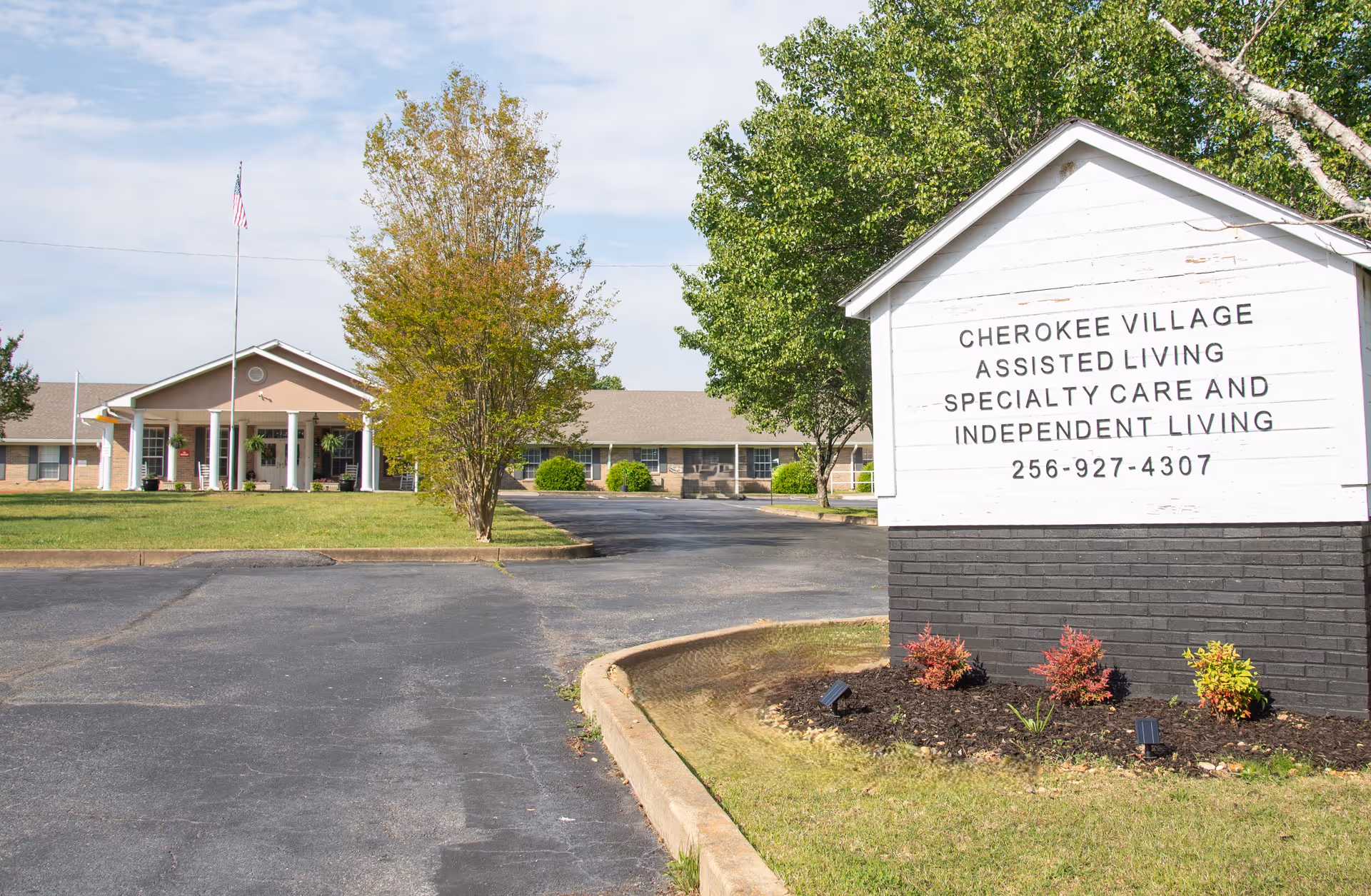 Front view of Cherokee Village Assisted Living Center showing the driveway, entrance building, and a white sign listing services and phone number.