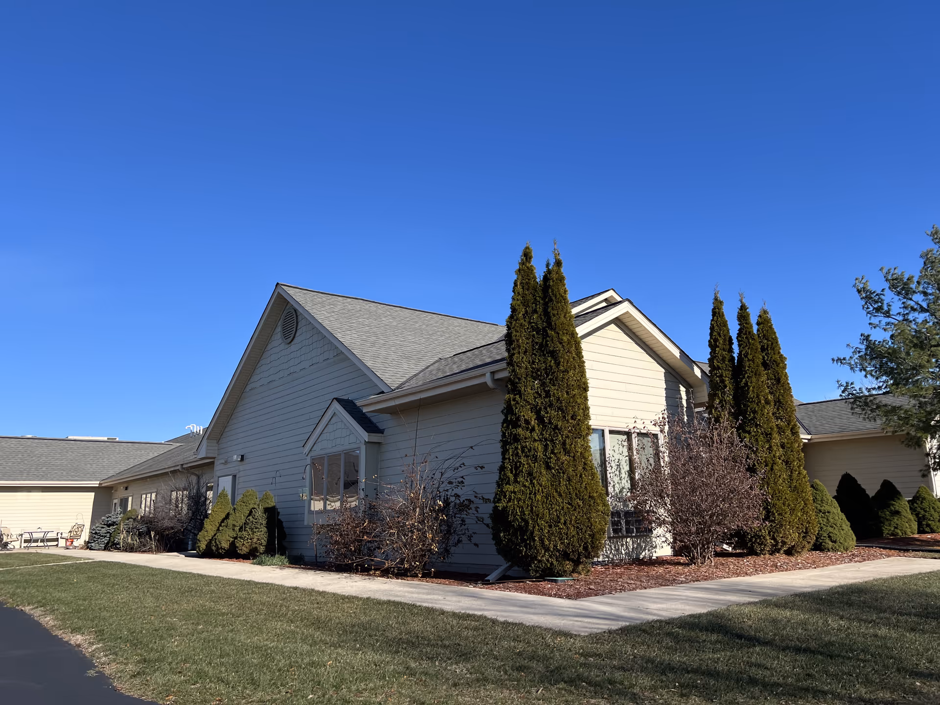Exterior view of a single-story residential building with beige siding and a gray roof under a clear blue sky. The building is surrounded by neatly trimmed bushes and tall evergreen trees, with a paved walkway and green lawn in the foreground.