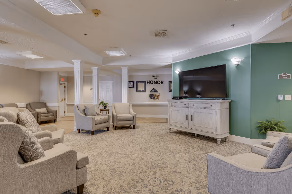 A spacious living room area in a senior living facility with multiple beige upholstered armchairs arranged around a large carpeted floor. A large flat-screen TV sits on a white wooden cabinet against a green accent wall. The wall behind the chairs features a 'Wall of Honor' display with framed items and a folded American flag. The room is softly lit with ceiling lights and wall sconces, and there are white columns and a small table with a plant in the background.