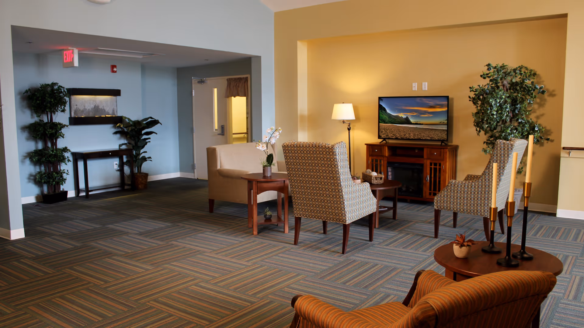 A cozy living room area in a senior living facility with patterned armchairs, a beige loveseat, a wooden TV stand with a flat-screen TV displaying a beach scene, a floor lamp, potted plants, and a small table with decorative candles. The walls are painted light yellow and blue, and the floor is covered with striped carpet tiles.