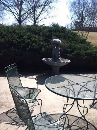 Outdoor patio area with a round metal table and two matching metal chairs on a concrete surface. In the background, there is a birdbath with a statue of two children holding an umbrella, surrounded by green bushes and leafless trees under a clear sky.