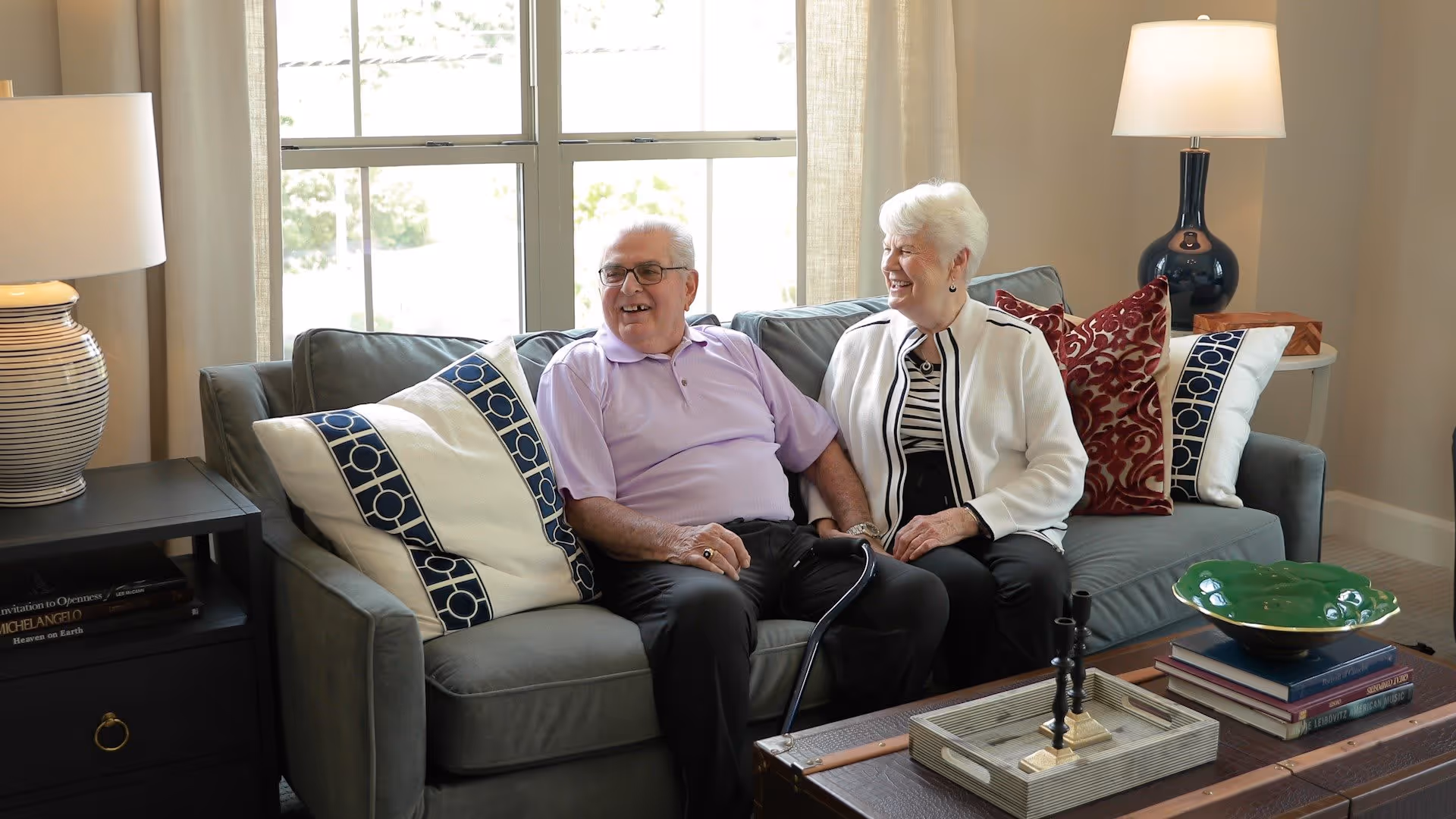 An elderly man and woman sitting together on a gray couch in a well-lit living room, smiling and holding hands. The room features decorative pillows, two table lamps on either side, a window with curtains behind them, and a wooden coffee table with books and decorative items.