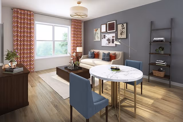 A modern living room with a beige sofa adorned with blue and orange pillows, a dark wooden coffee table, a round white marble dining table with two blue chairs, a wooden shelving unit with books and plants, and large windows with patterned orange and white curtains letting in natural light.
