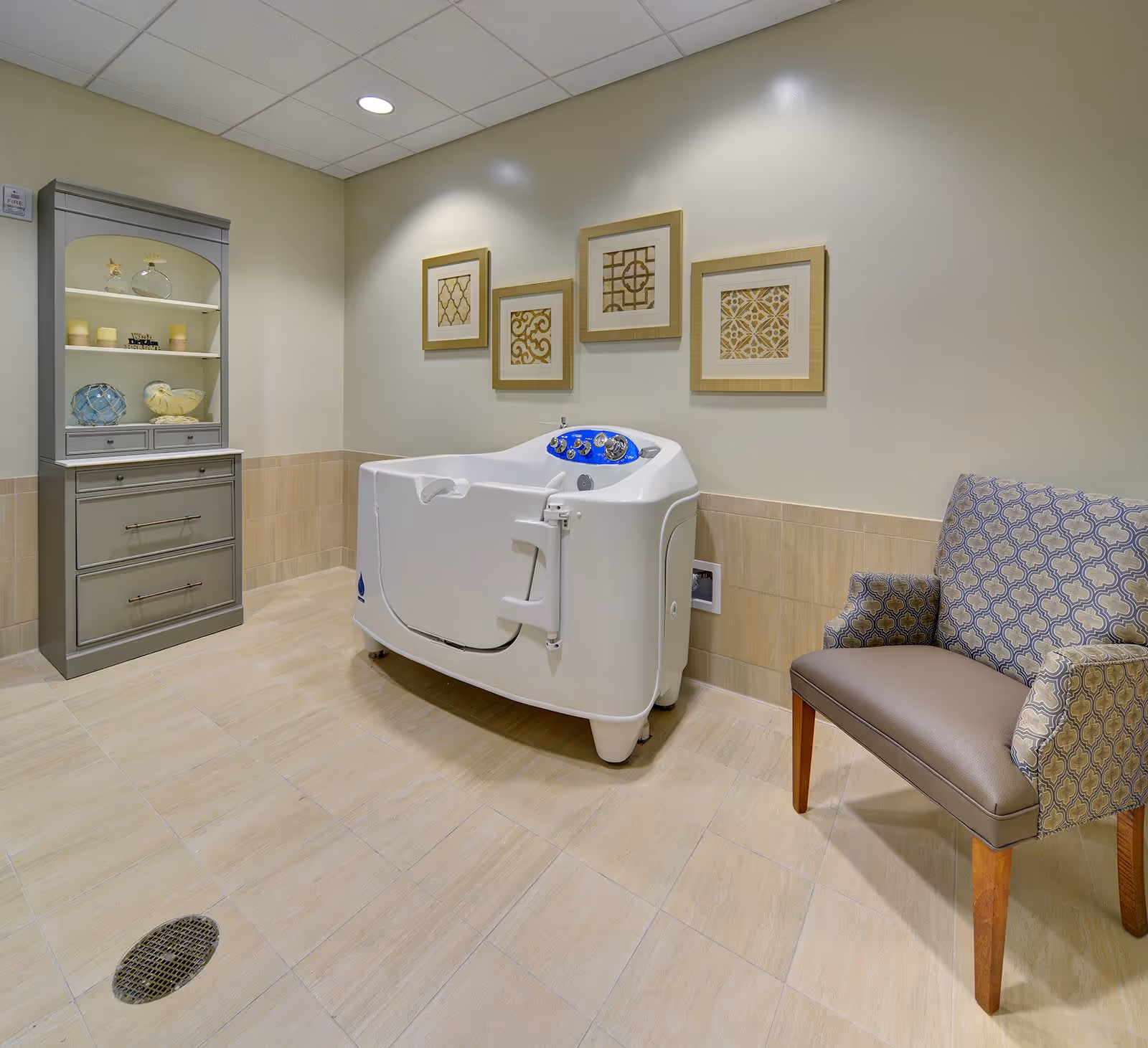 A clean, well-lit bathing room featuring a white walk-in bathtub with blue controls, a patterned armchair with wooden legs, a gray cabinet with decorative items, and four framed decorative wall art pieces above the bathtub.