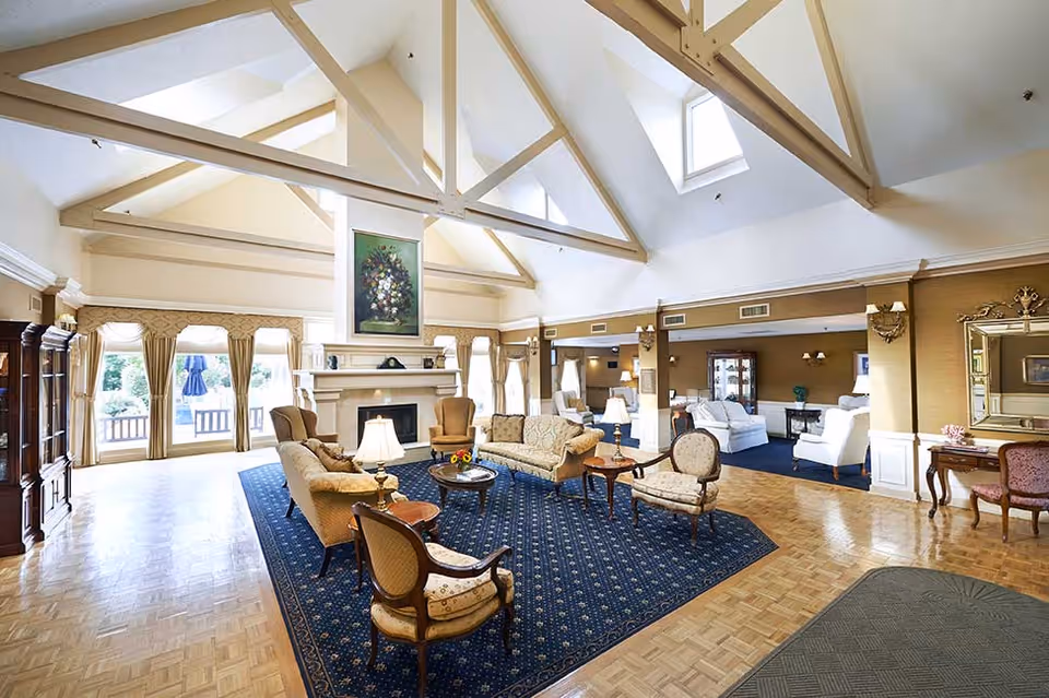 Spacious and well-lit living room area with high vaulted ceilings featuring exposed beams. The room includes multiple seating arrangements with sofas, armchairs, and side tables on a large blue patterned rug. There is a fireplace with a floral painting above it, large windows with curtains letting in natural light, and wooden parquet flooring. The room has a classic and elegant decor style.
