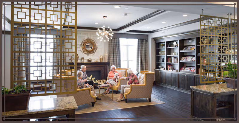 A cozy living room area in a senior living facility with four elderly people sitting on yellow armchairs around a coffee table. The room features large windows with curtains, a fireplace with decorative items, a sunburst mirror above the fireplace, and built-in bookshelves filled with books and magazines. Decorative gold geometric room dividers partially frame the space.