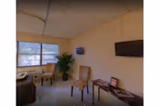 A small room with beige walls and a tiled ceiling, featuring two beige chairs, a small table with magazines and a framed photo, a potted plant near a window with blinds, and a wall-mounted flat screen TV.