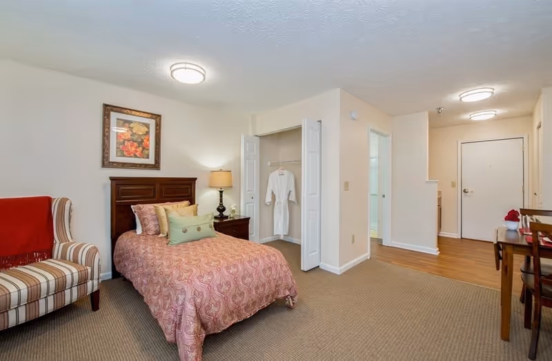 Interior of a senior living studio apartment showing a bed with patterned bedding, nightstand and lamp, striped armchair, open closet with robes, and the entryway with a small table.