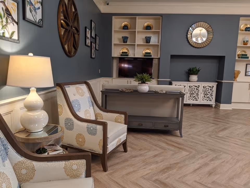 Comfortable seating area with patterned armchairs, a lamp, console table, shelving and wall decor in a senior living lounge.