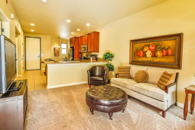 A cozy living room area with a beige sofa adorned with three decorative pillows, a round brown leather ottoman, and a dark brown armchair. A large framed painting of colorful flowers hangs on the wall above the sofa. The room has beige carpet flooring and opens into a kitchen area with wooden cabinets, granite countertops, and stainless steel appliances. A TV is placed on a stand to the left side of the image.