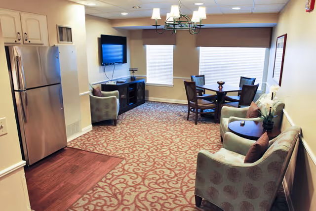 Carpeted common living area with armchairs, a round dining table, a wall-mounted TV, and a stainless steel refrigerator.