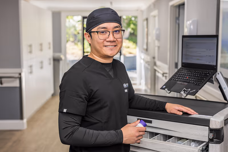 A smiling healthcare worker wearing black scrubs and a black cap stands next to a medical cart with a laptop on top in a bright hallway of a care facility.