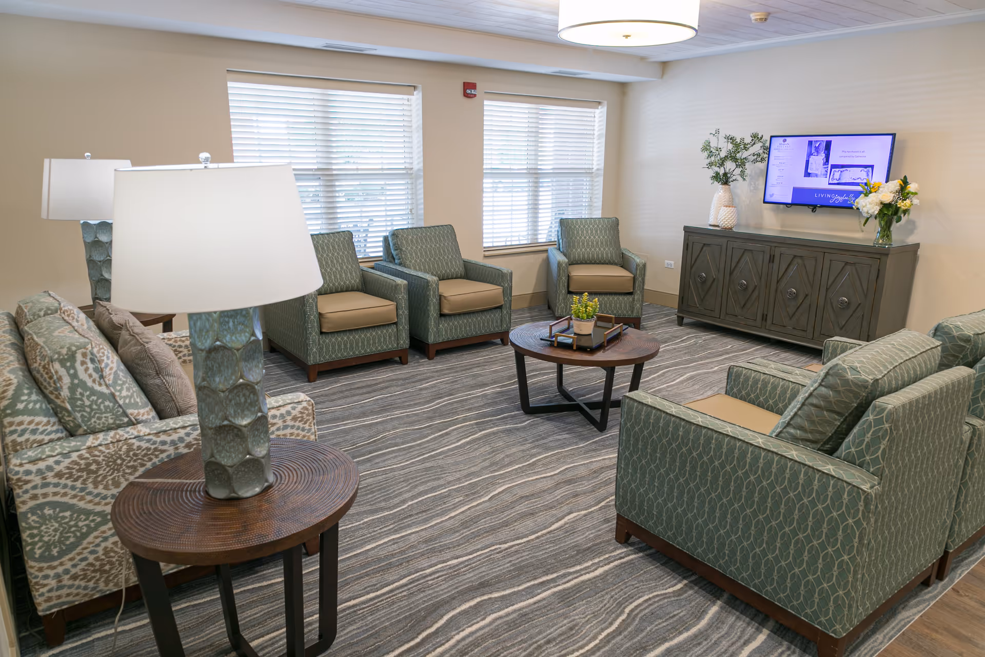 A cozy living room with patterned armchairs and a sofa arranged around a round wooden coffee table. The room features a gray and white striped carpet, two large windows with blinds, a side table with a lamp, a cabinet with decorative vases and flowers, and a wall-mounted TV.