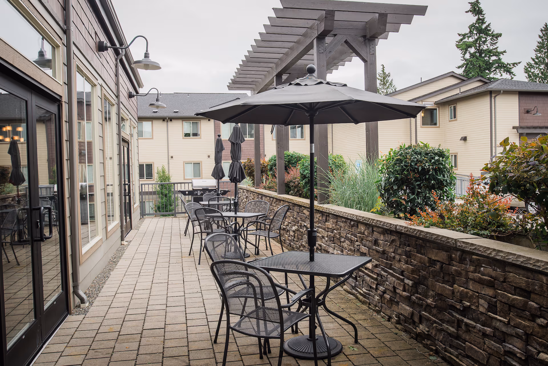 Outdoor patio with metal tables and chairs, umbrellas, a pergola, and landscaping beside a stone half-wall and building.