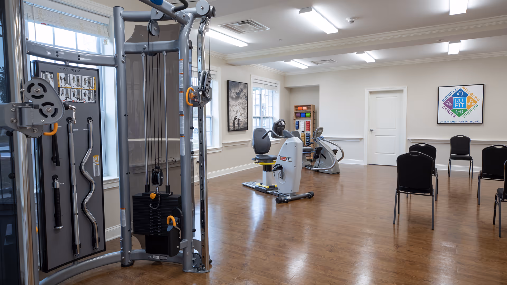 A fitness room with exercise equipment including a multi-station weight machine, two stationary bikes, and a rack of colorful dumbbells. The room has wooden flooring, white walls, large windows, and several black chairs arranged along the wall. A framed poster about fitness is displayed on the wall.