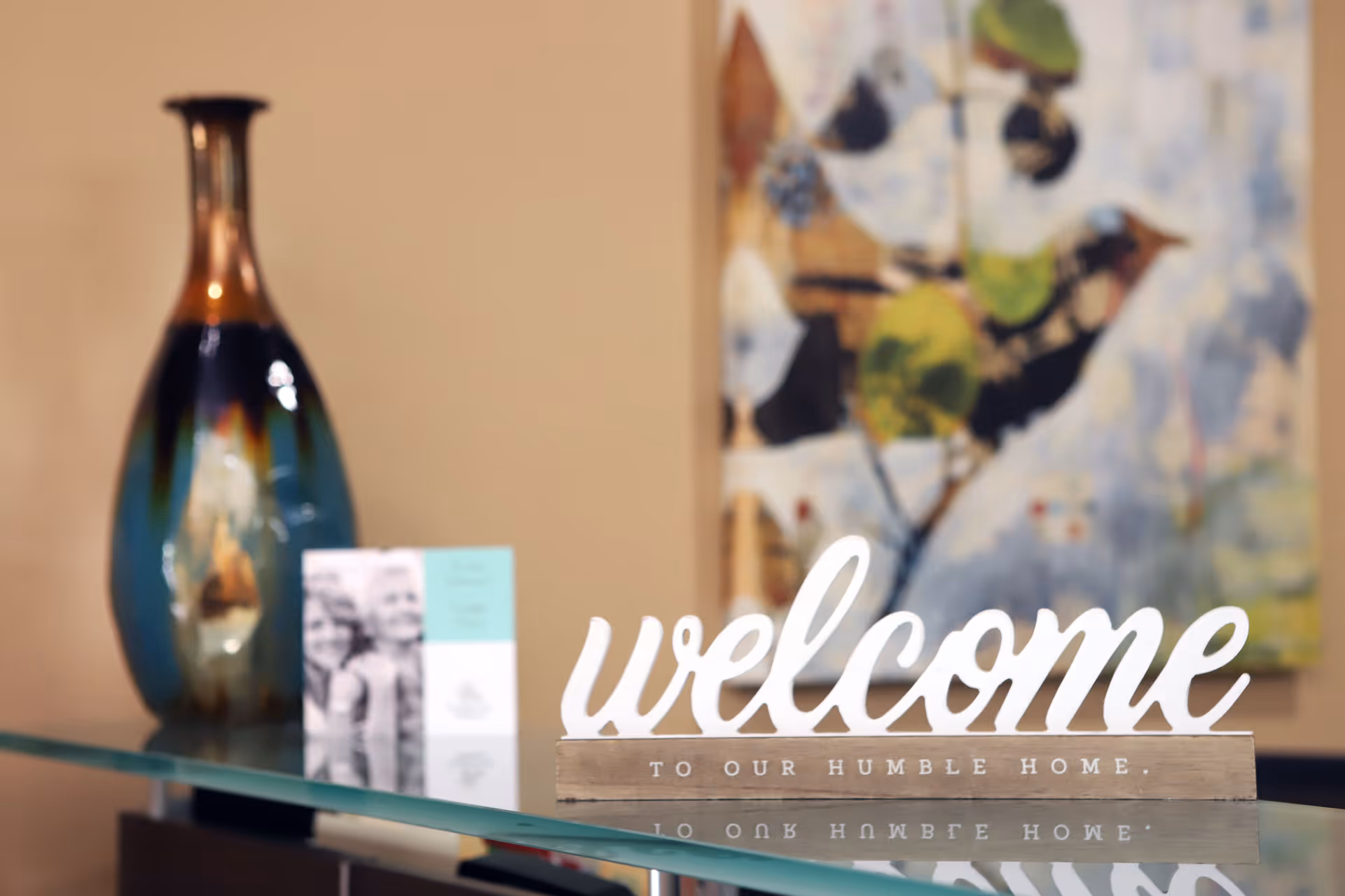 A decorative glass table with a large blue and brown vase, a small card, and a wooden sign that says 'welcome to our humble home' in white letters. In the background, there is a colorful abstract painting on the wall.