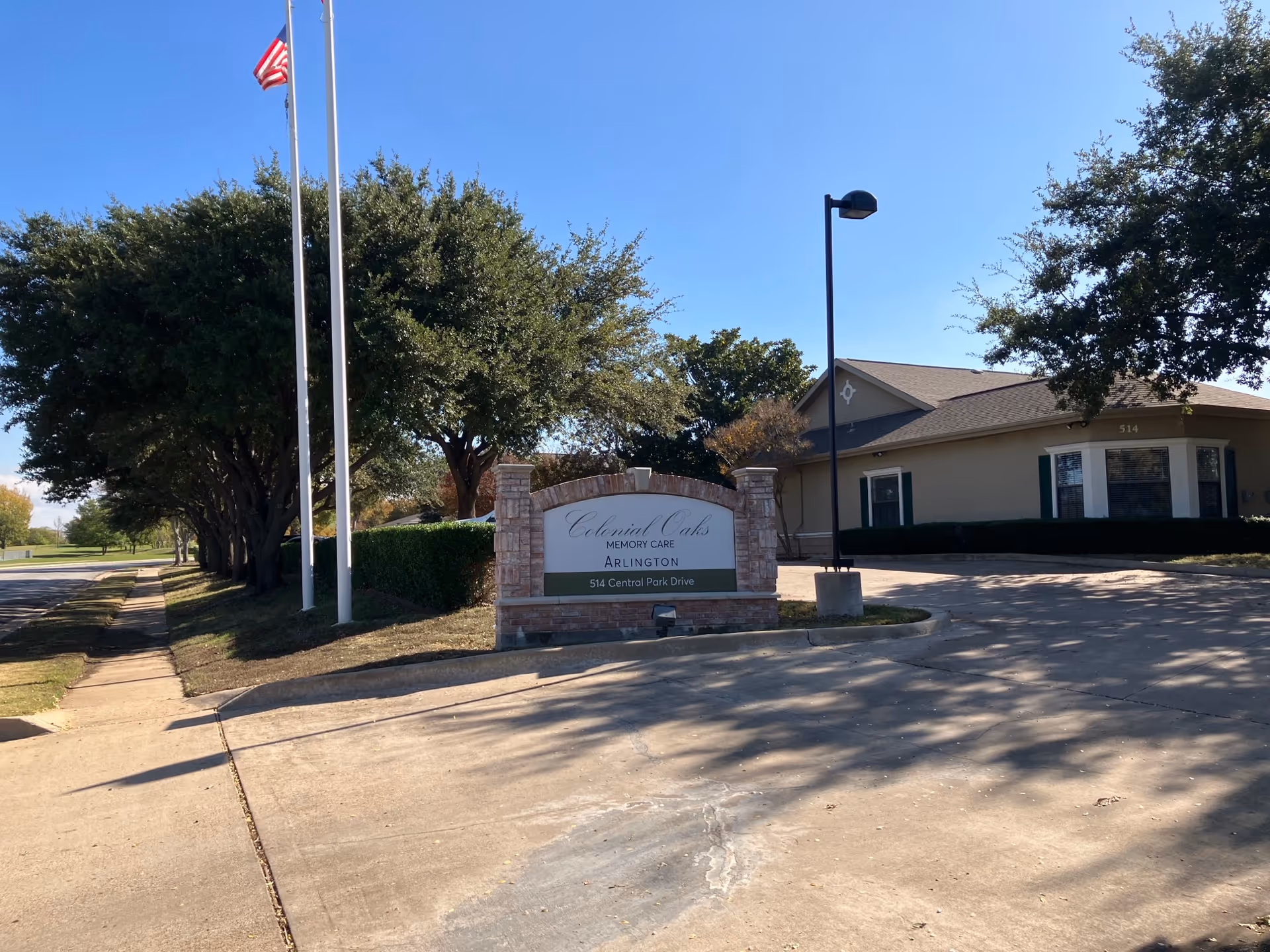 Exterior view of Colonial Oaks Memory Care Arlington facility with a brick sign displaying the name and address 514 Central Park Drive. There are two flagpoles with an American flag, trees lining the sidewalk, and a single-story building in the background under a clear blue sky.