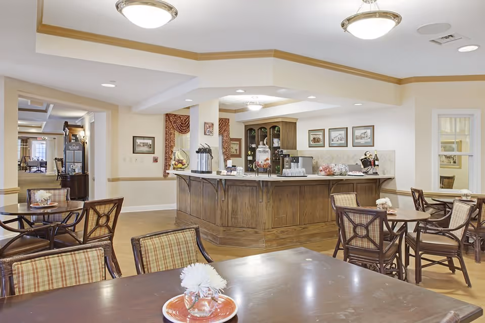 A bright and welcoming dining area in a senior living facility featuring several wooden tables and chairs with plaid cushions. In the background, there is a wooden counter with coffee dispensers, a water dispenser, and decorative items. The walls are adorned with framed pictures and the room has warm lighting from ceiling fixtures.