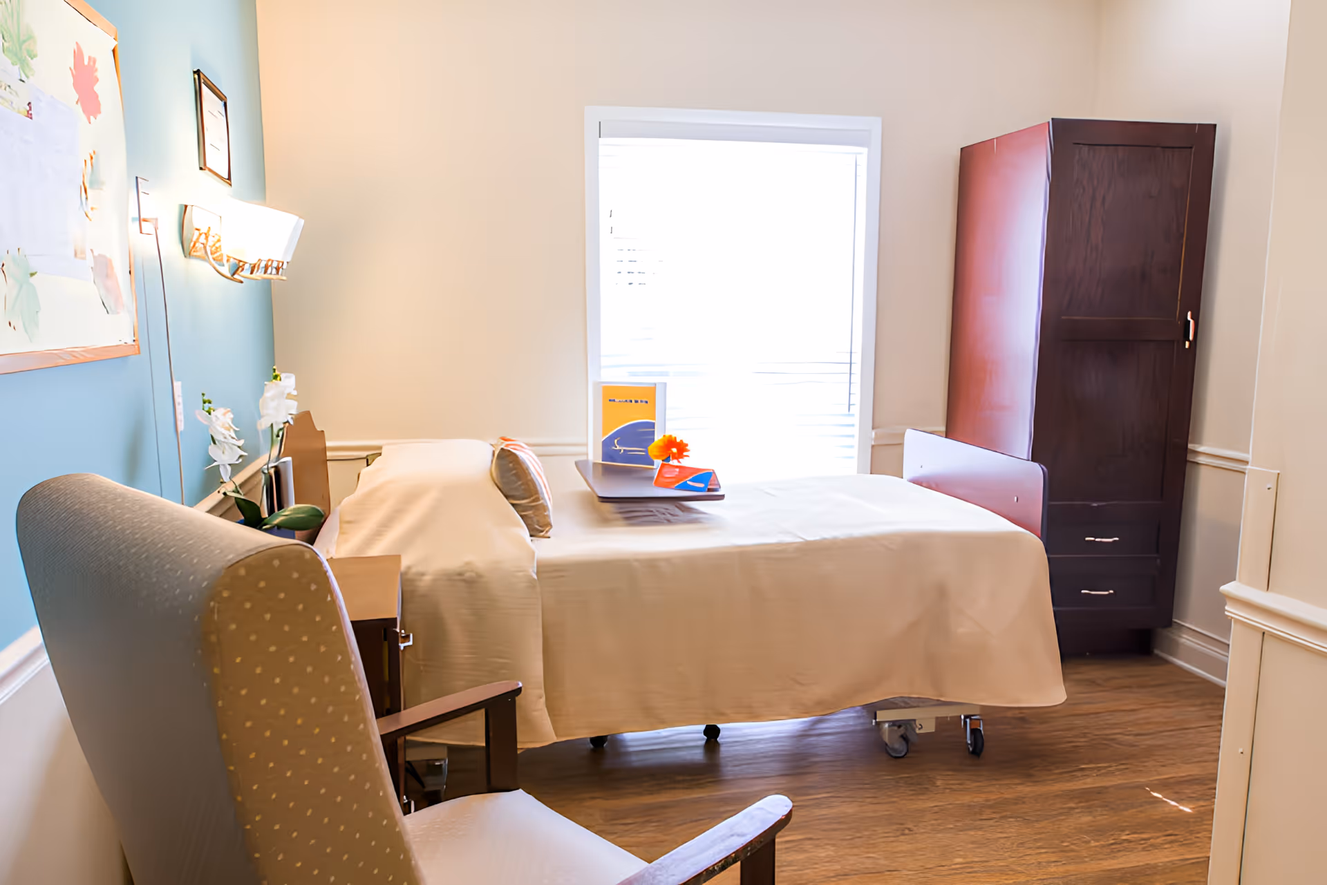 A bright nursing center bedroom with a single bed covered in white linens, a wooden bedside table with a flower and some books on a tray, a cushioned armchair with wooden armrests, a tall dark wooden wardrobe, and a window with blinds letting in natural light.