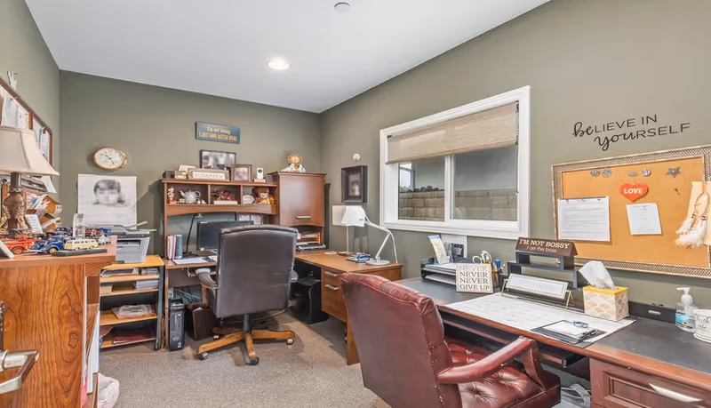 Small private office with two desks, leather chairs, shelving, a window, and a cork bulletin board.