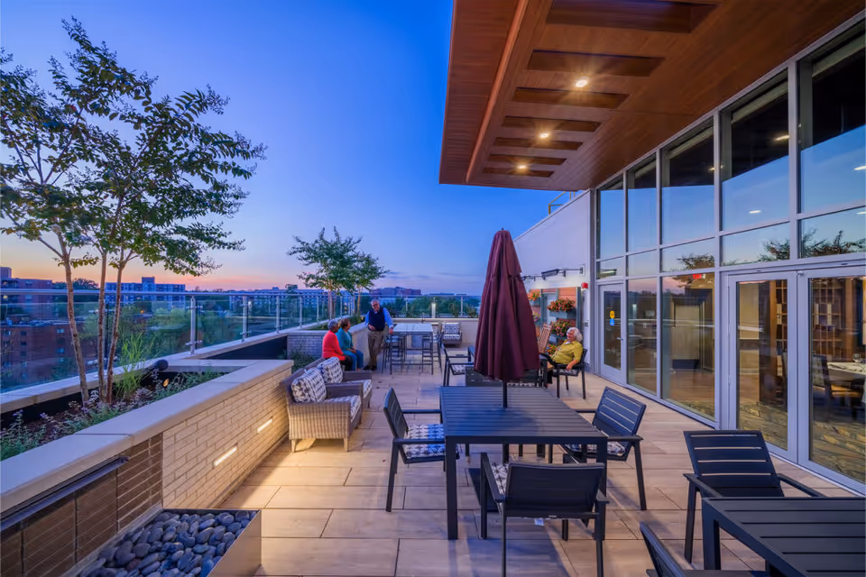 Outdoor patio area at sunset with several people sitting and chatting. The space features modern outdoor furniture including tables, chairs, and cushioned seating. There are small trees planted along a glass railing overlooking a cityscape. The building has large glass windows and a wooden ceiling with recessed lighting.