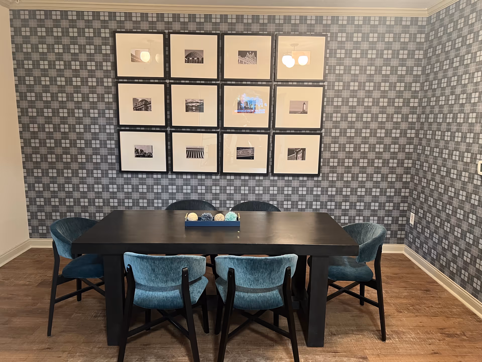 Dark dining table with six teal upholstered chairs against a gray patterned wallpapered wall featuring a grid of framed black-and-white photos.