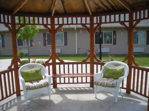 View from inside a wooden gazebo with a bench and two white wicker chairs with colorful cushions, overlooking a grassy area and a building with windows and shutters in the background.