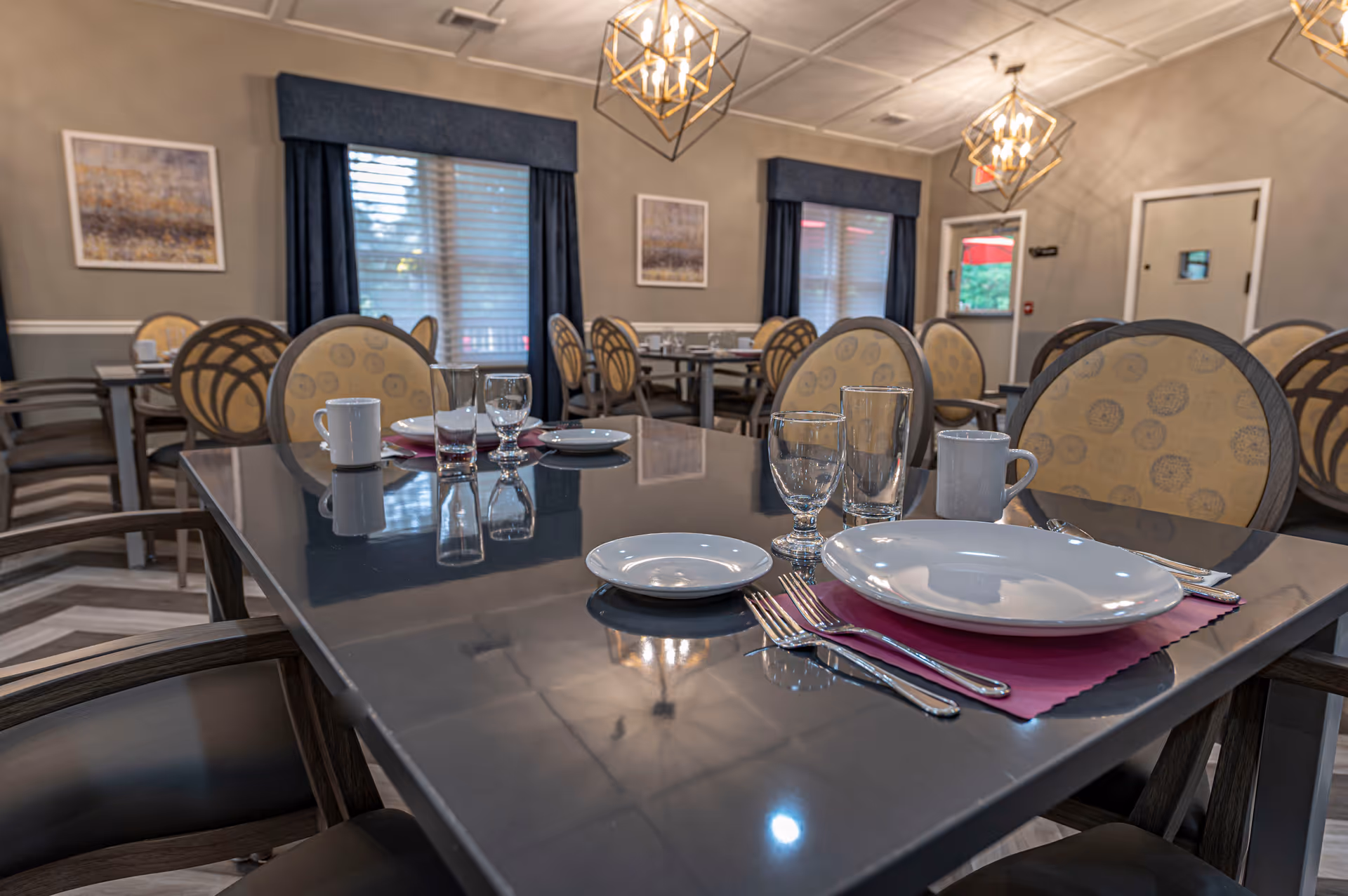 Dining room with neatly set tables, plates, glasses, and upholstered round-backed chairs under decorative lighting.