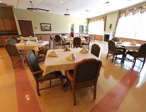 A dining hall with round tables set with napkins, chairs, large windows, and plants.