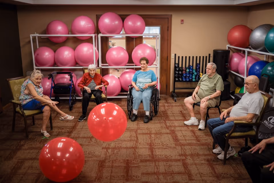 A group of elderly people sitting in a circle in a room with exercise equipment, including large pink exercise balls and dumbbells. Two large red exercise balls are on the floor in the center of the circle. The room has carpeted flooring and beige walls.