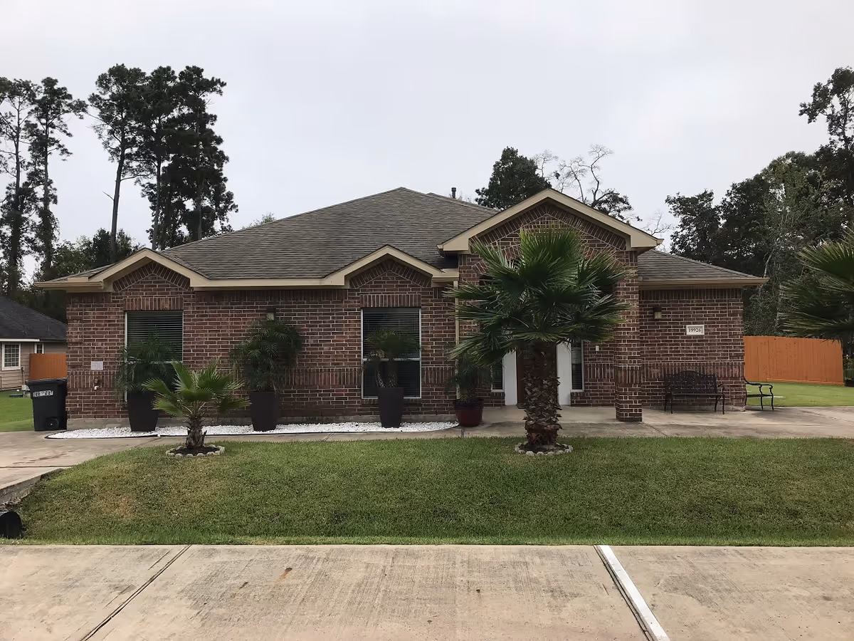 Front exterior view of a single-story brick building with a gray shingled roof, several windows, and a small porch area. The lawn is neatly maintained with small palm trees and potted plants near the entrance. There is a concrete driveway and sidewalk in front of the building, and tall trees are visible in the background.