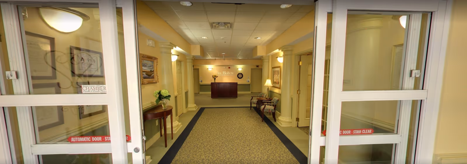 View through automatic glass doors into a well-lit hallway of Fox Hollow Senior Living Community. The hallway features beige walls with white columns, framed artwork, a small table with a vase of flowers on the left, and two upholstered chairs with a lamp on the right. At the end of the hallway is a dark wood reception desk with the Fox Hollow Senior Living Community sign on the wall behind it.