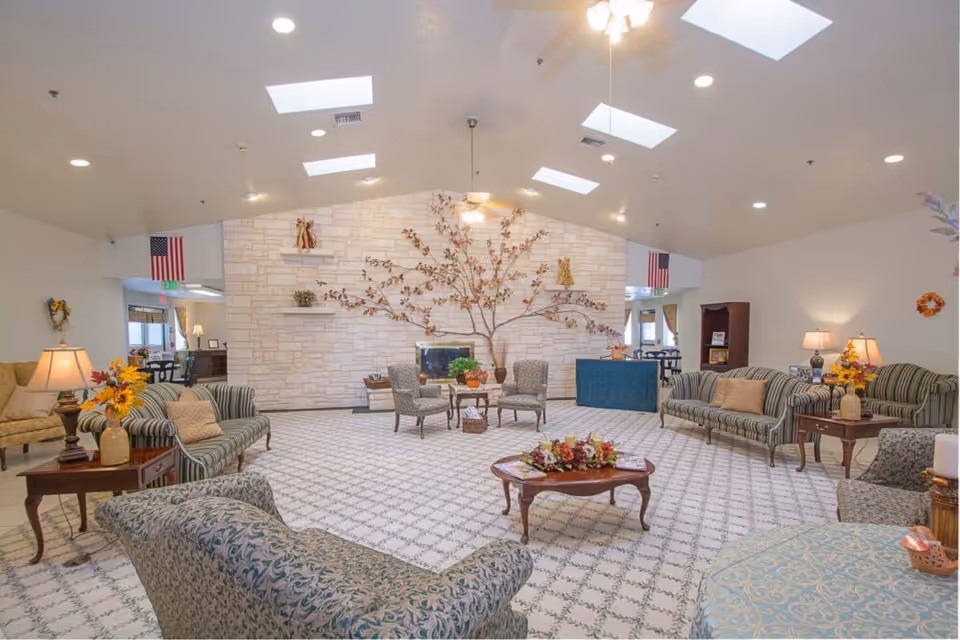A spacious and well-lit living room area in a senior living facility with patterned carpet flooring, several upholstered sofas and armchairs arranged around a wooden coffee table with a floral centerpiece. The back wall features a stone fireplace with a decorative metal tree sculpture above it. The room has skylights, ceiling fans, table lamps, and American flags on either side of the fireplace.