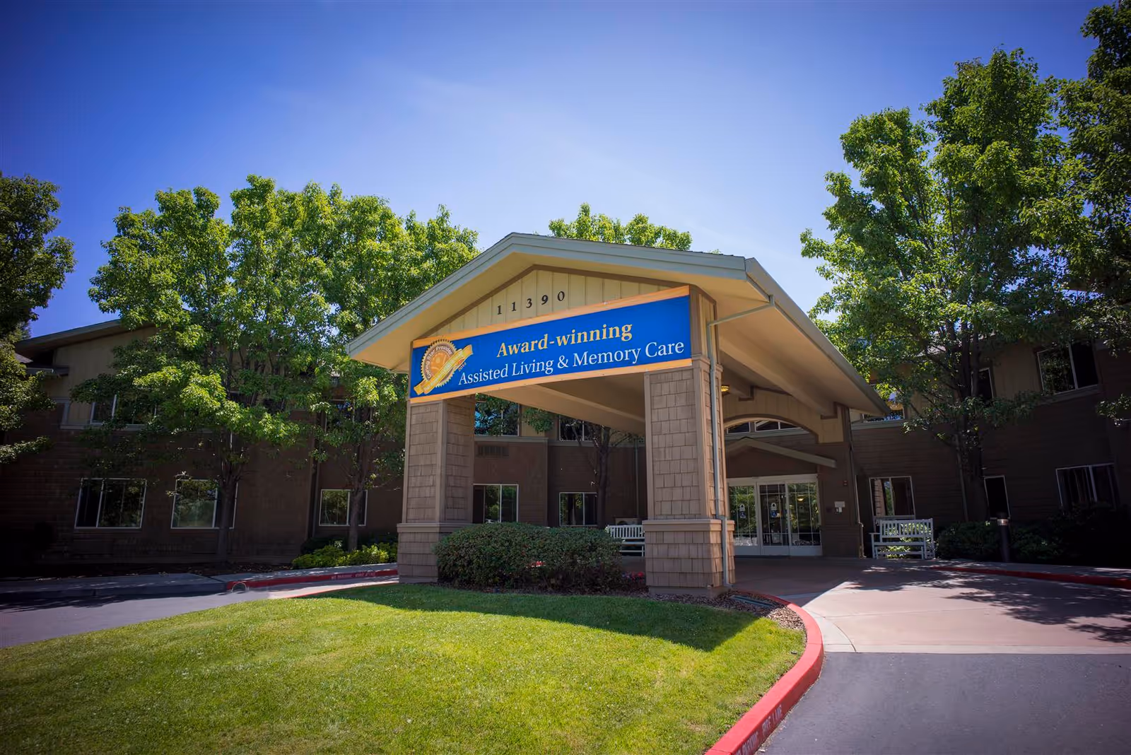 Entrance canopy of a two-story assisted living facility with a sign reading 'Award-winning Assisted Living & Memory Care' and trees around the driveway.