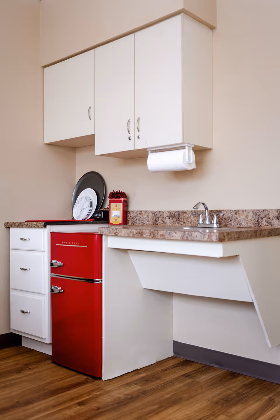 A small kitchen area with white cabinets, a granite countertop, a stainless steel sink with a faucet, a red mini refrigerator, and a paper towel holder mounted under the upper cabinets. There are plates and a container on the countertop, and the floor is wood.