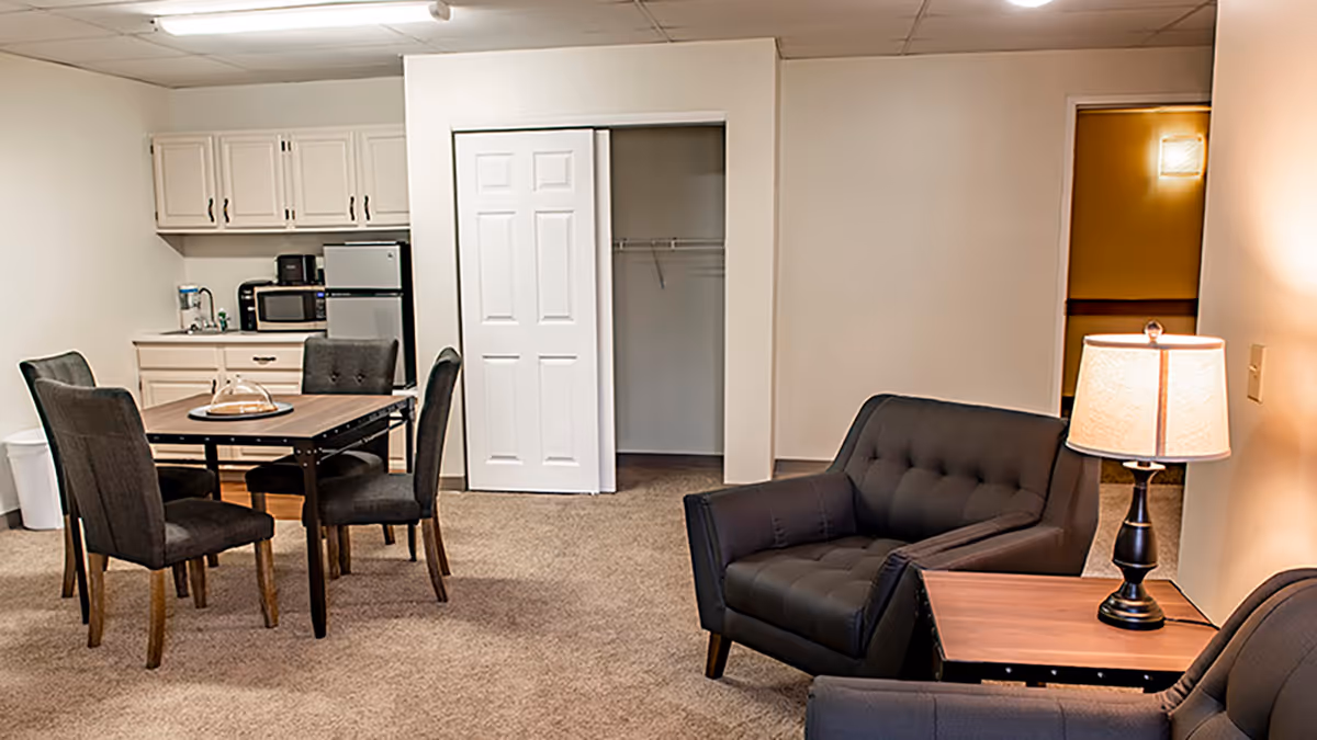 Interior view of a senior living facility room featuring a small kitchenette with white cabinets, a microwave, and a mini refrigerator. In front of the kitchenette is a dining table with four dark upholstered chairs. To the right, there are two dark armchairs with a wooden side table and a lit table lamp. The room has beige carpet and white walls, with an open closet and a doorway visible in the background.