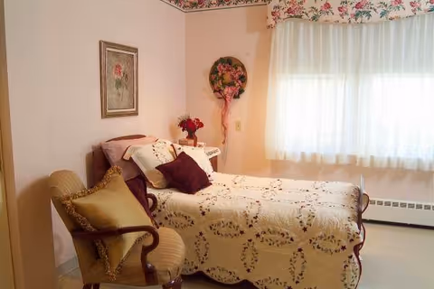 A cozy single bedroom with a floral quilted bed, decorative pillows, an upholstered armchair, wall decor, and a curtained window.