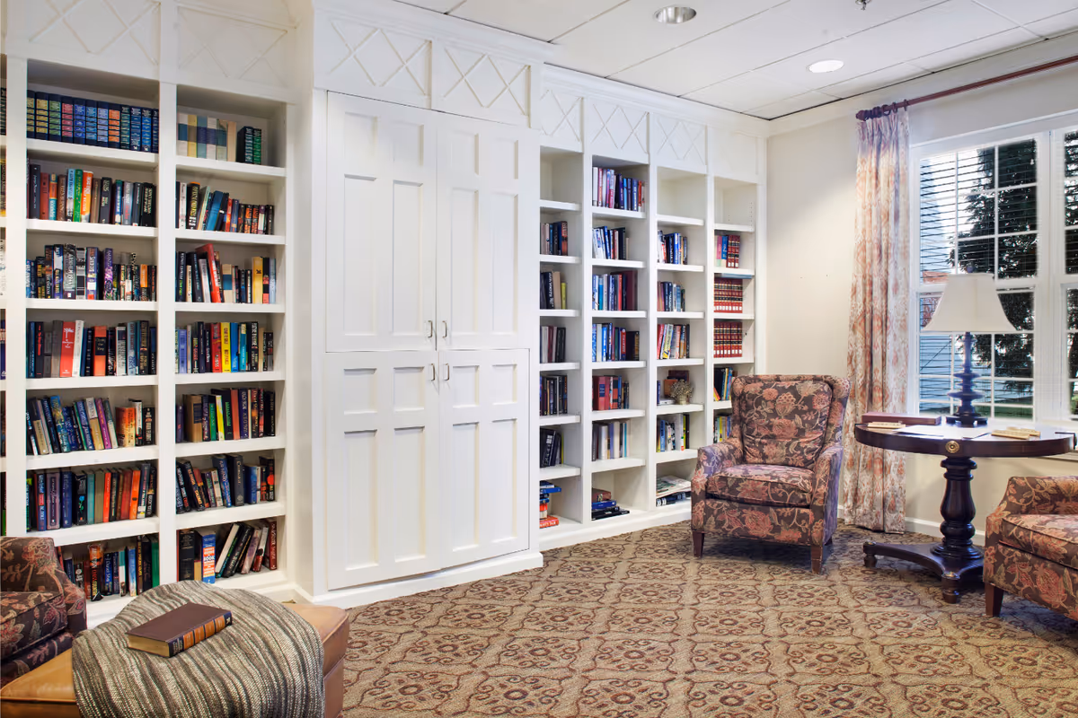 Cozy reading room with built-in white bookshelves, patterned armchairs, a round side table with a lamp, and a window with curtains.