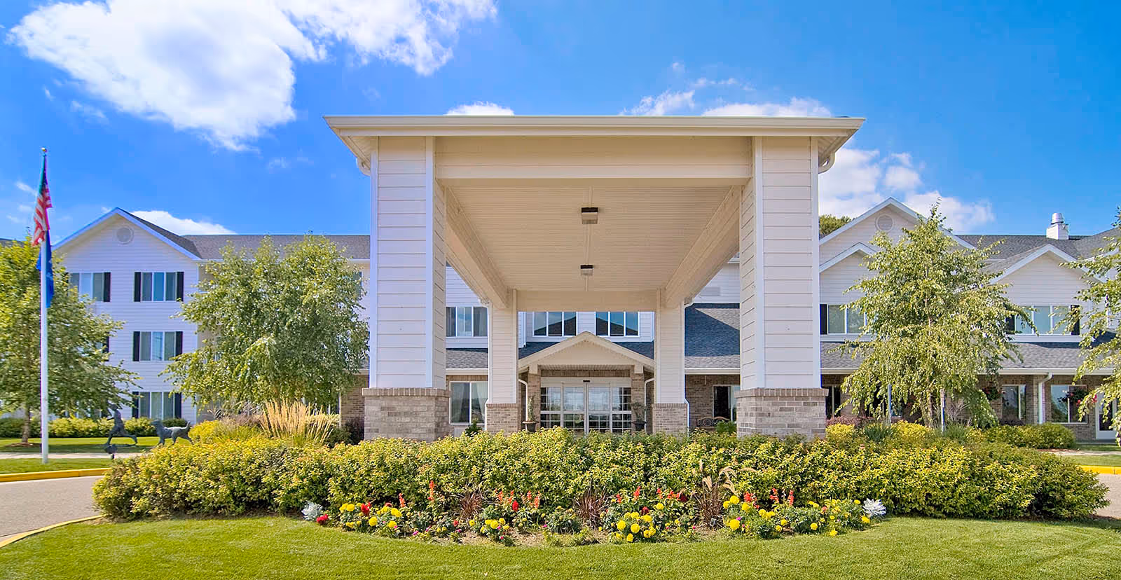 Front exterior view of Oakwood Hills - Sky Active Living facility with a covered entrance, well-maintained landscaping including green bushes and colorful flowers, and a clear blue sky with some clouds.